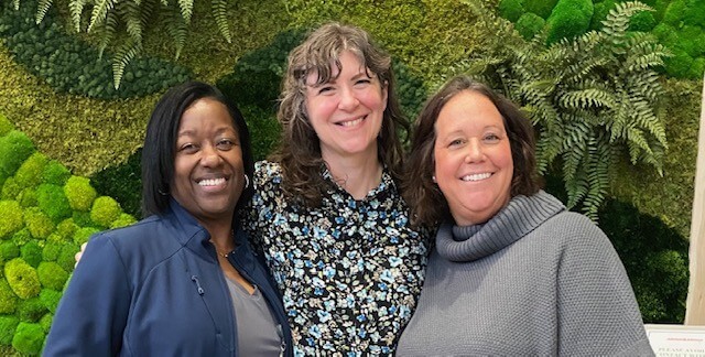 Three women from the shoulders up, posing in front of a plant wall